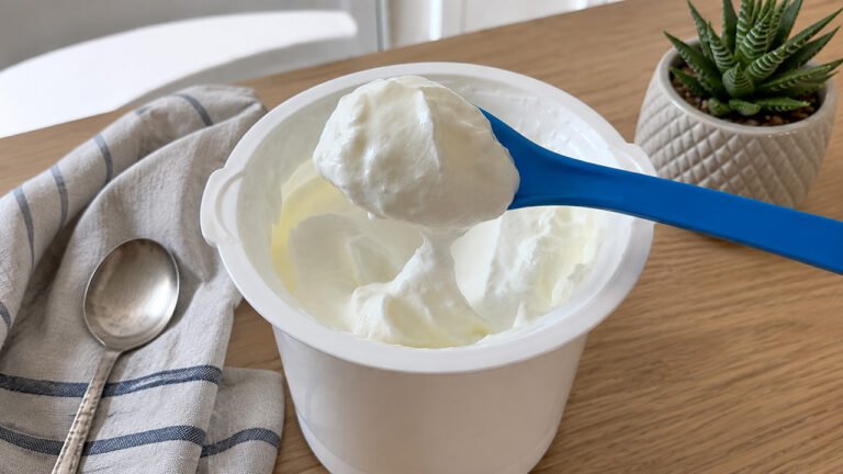 Yogurt greco **denso e cremoso** appena colato, sollevato con una spatola blu nel contenitore della yogurtiera. Sullo sfondo, strofinaccio, cucchiaio e piantina grassa.