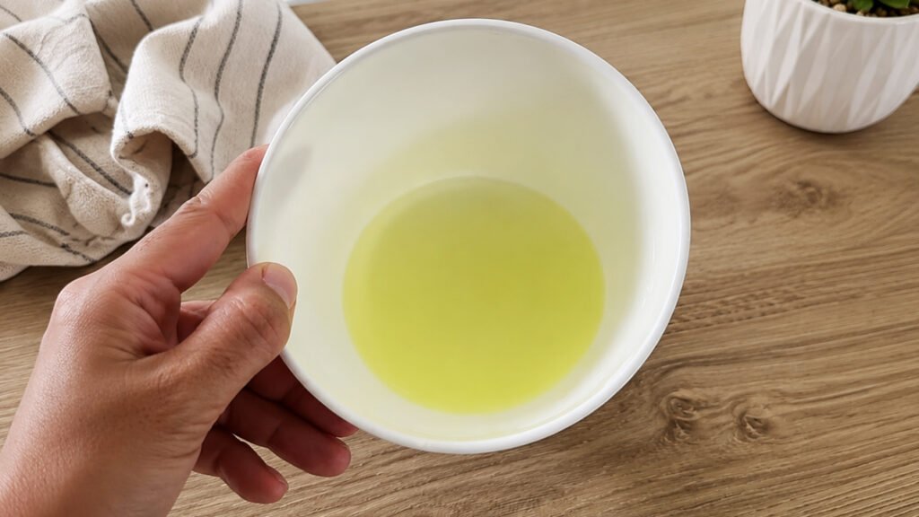 Una ciotola bianca tenuta in mano con il siero giallo della colatura. Sullo sfondo ci sono uno strofinaccio e una piccola pianta grassa.