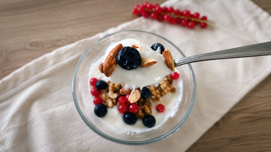 Primo piano dello yogurt greco cremoso servito in una ciotola di vetro, con mandorle, noci, mirtilli e ribes, mentre una cucchiaiata viene sollevata con il cucchiaio.